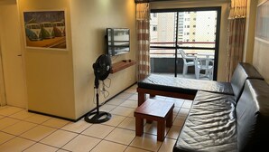 A small living room with a sofa, a TV, a fan, and a small table. There is a balcony with a view of buildings. - Porto de Iracema Apartamentos (Fortaleza)