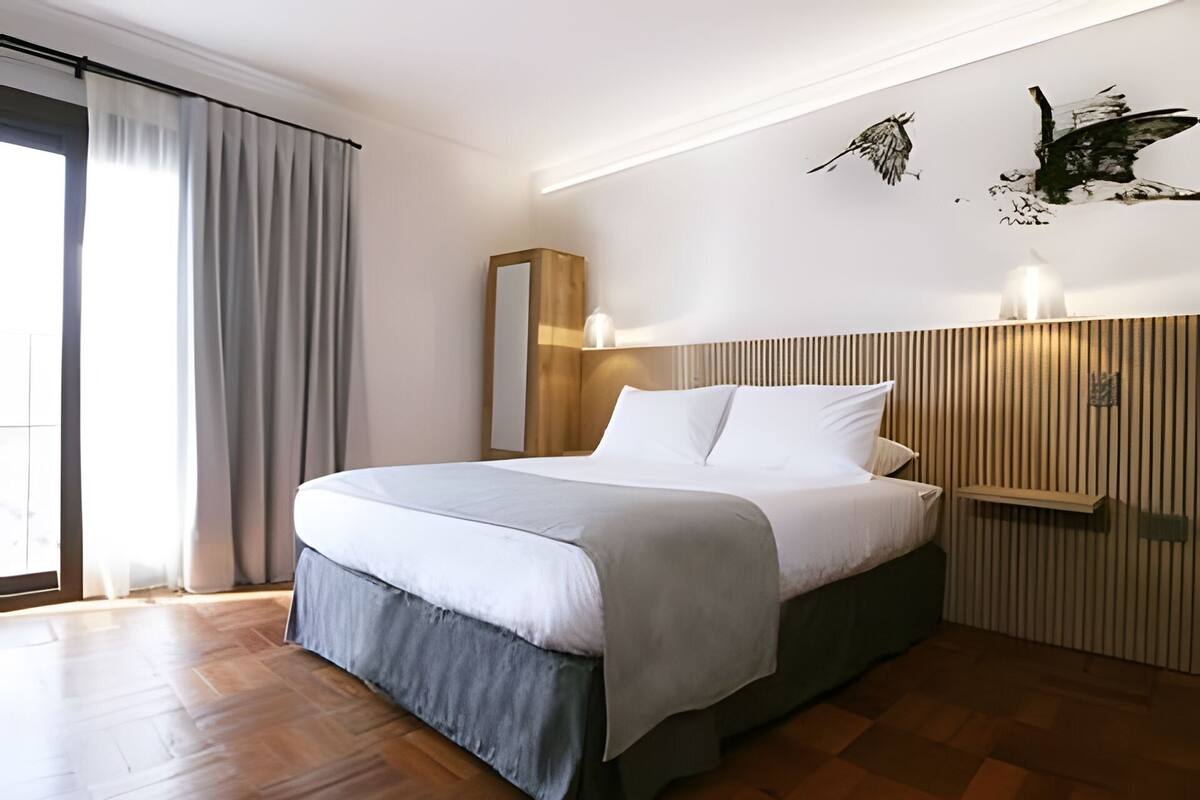 A modern hotel room with a large bed, a wooden headboard, a mirror, and wall-mounted lights.