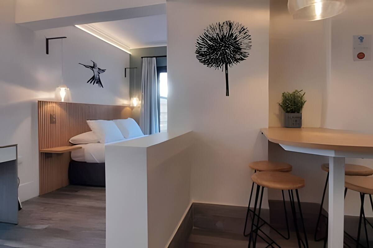 A modern hotel room with a bed, bedside table, and a small dining area with stools.