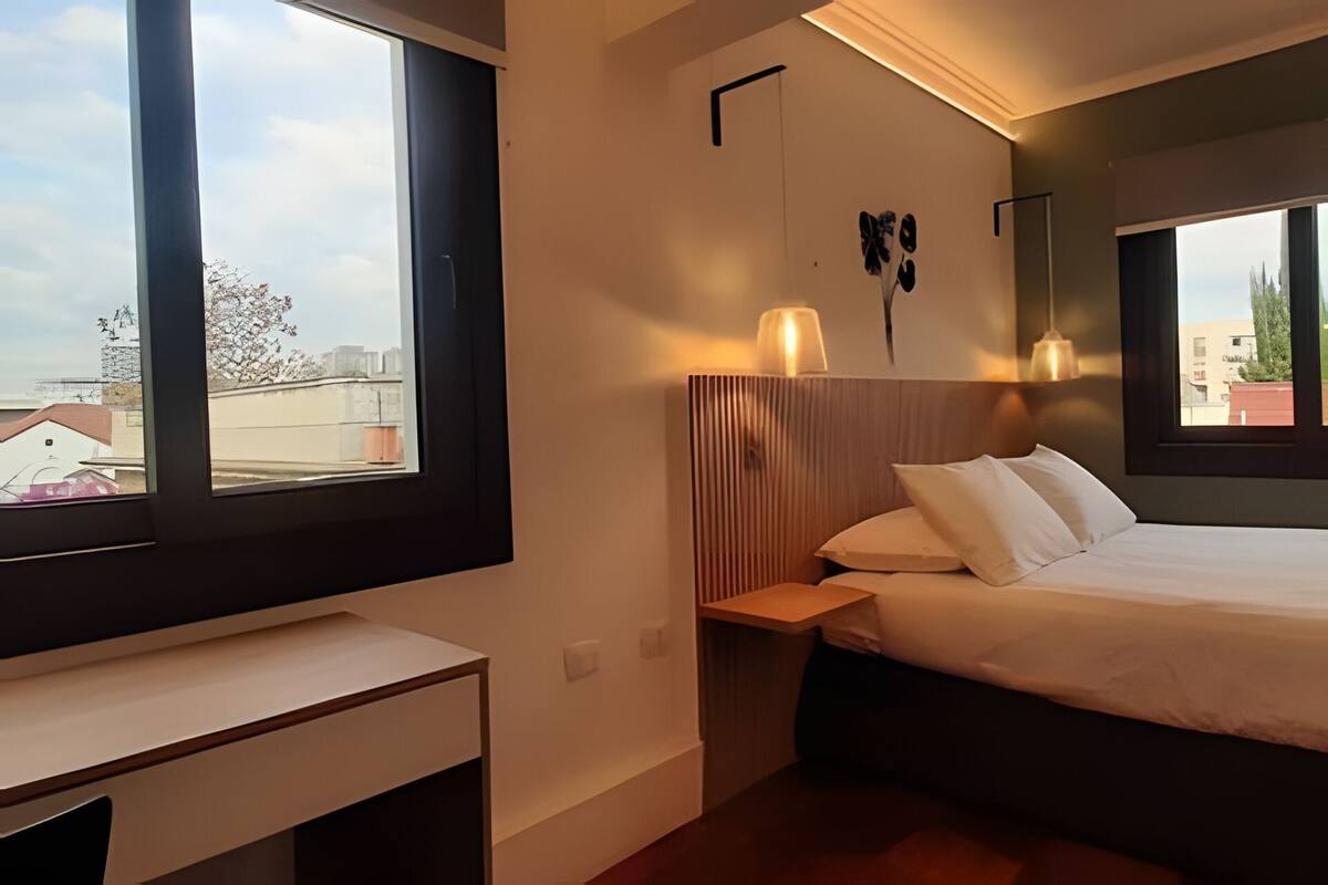 A bedroom with a bed, bedside table, and a view of the cityscape through large windows.