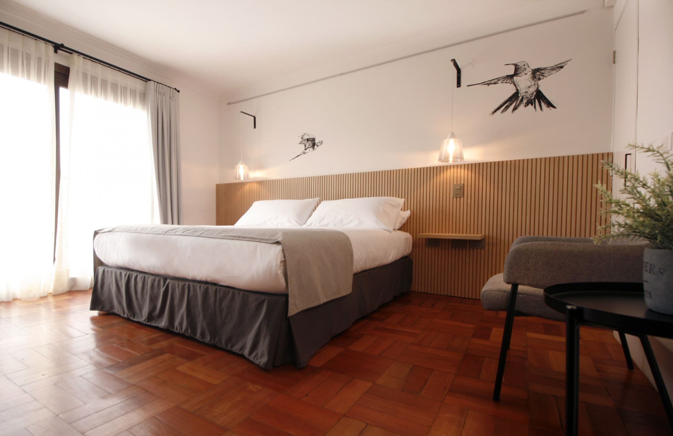 A modern bedroom with a wooden floor, a bed with a grey skirt, a bedside table, and a wall-mounted light fixture.