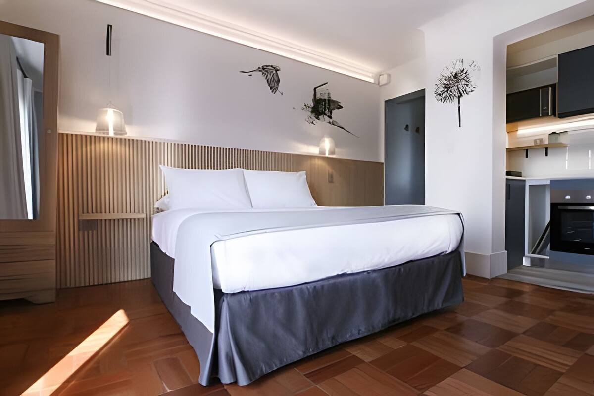A modern hotel room with a large bed, wooden headboard, and a built-in kitchen area.
