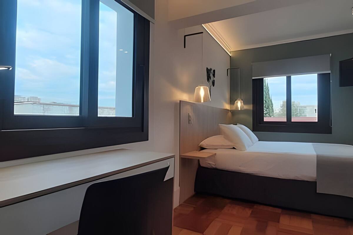 A compact hotel room with a desk, a bed, and a large window offering a view of the cityscape.