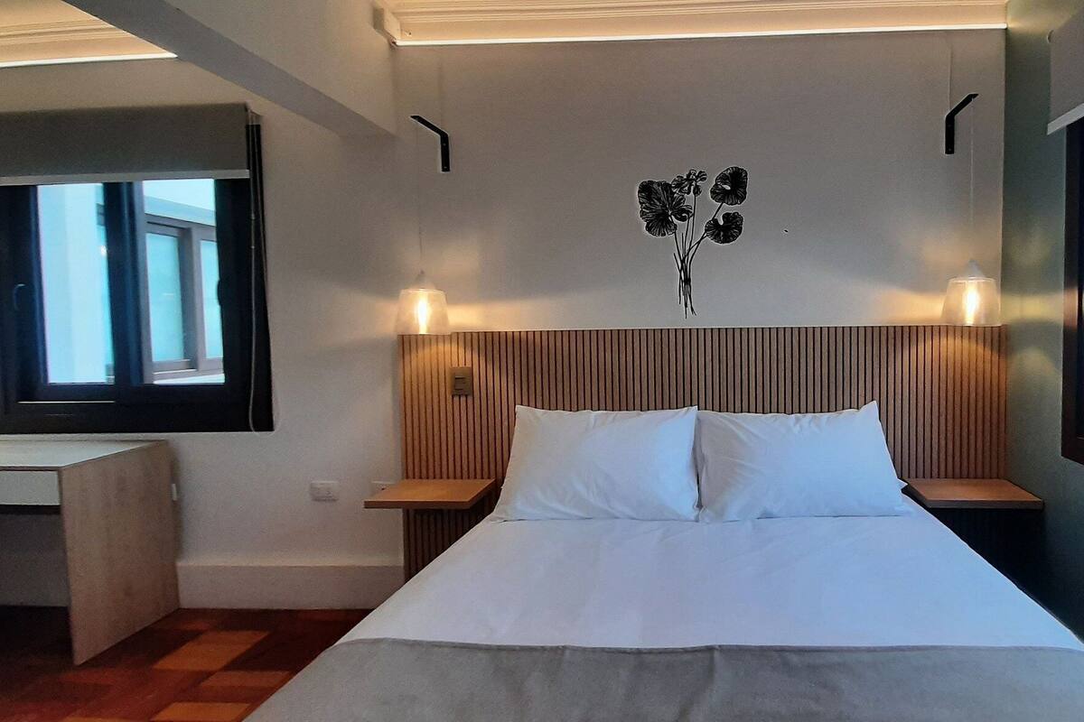 A hotel room with a bed, a desk, a chair, a window, and a wall-mounted light fixture.