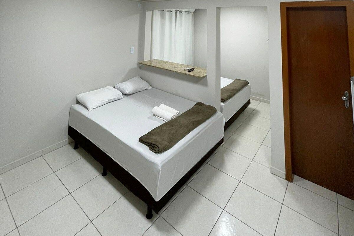 Una cama individual con ropa de cama blanca, cabecera de madera, una pequeña estantería encima y una puerta a la derecha.