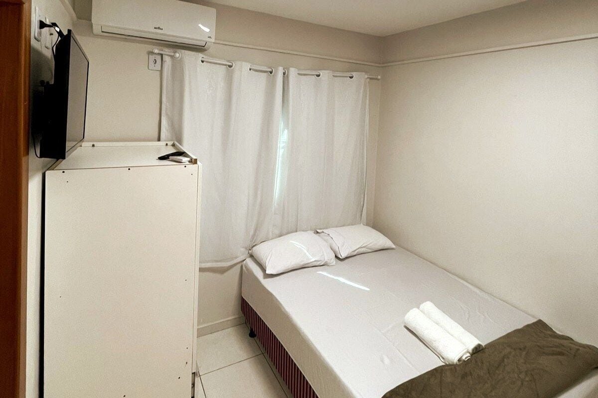 A small, single-bed room with a white bed, a white TV on a stand, and a white air conditioning unit above the bed.