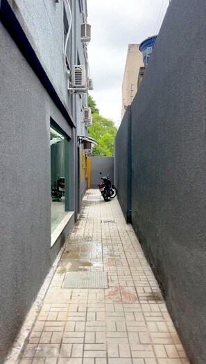 Un estrecho callejón con una motocicleta estacionada a la derecha, flanqueado por edificios con unidades de aire acondicionado y un ventanal visible a la izquierda.