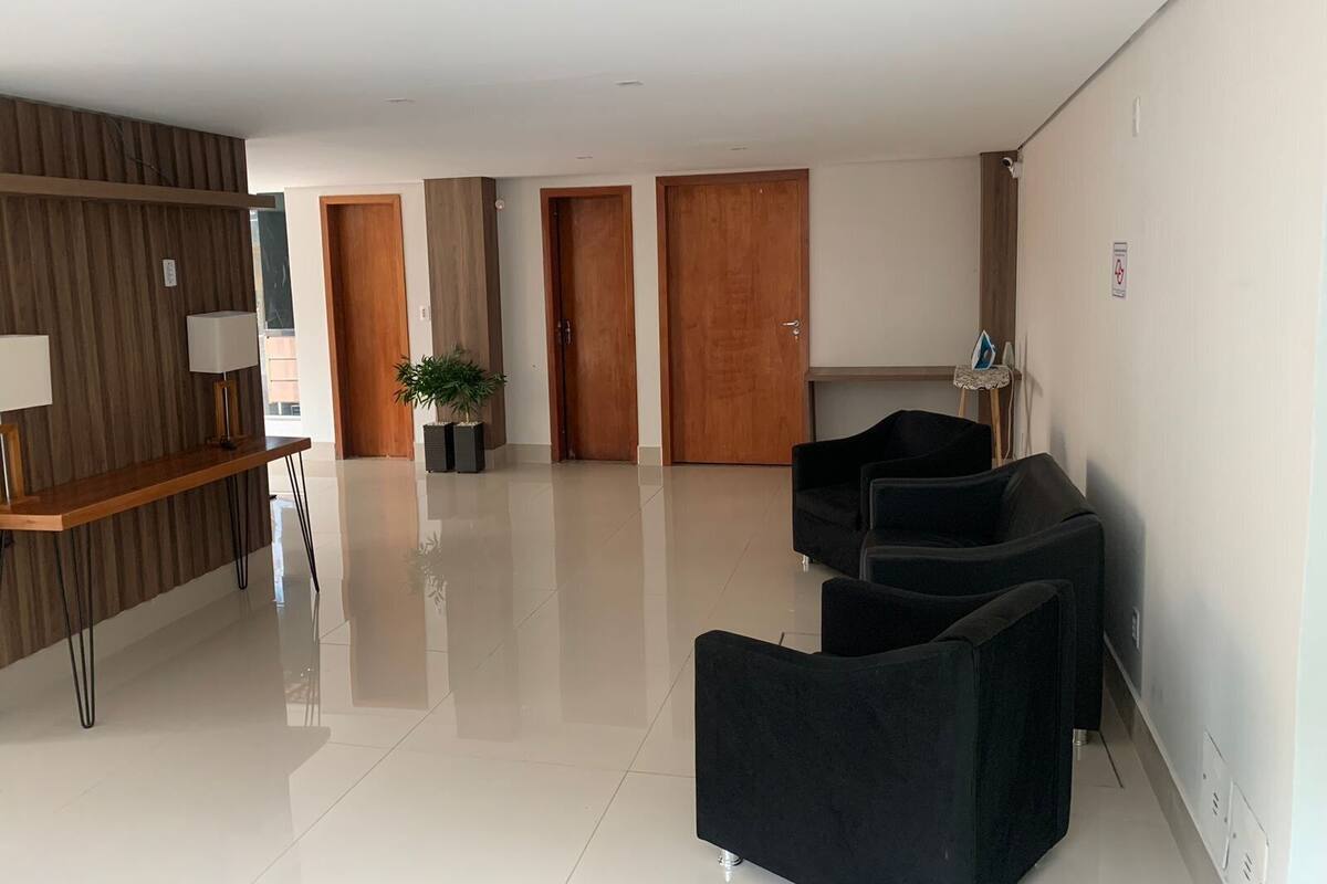 Um saguão de hotel moderno com balcão de recepção, assentos pretos, portas de madeira e uma planta em vaso.