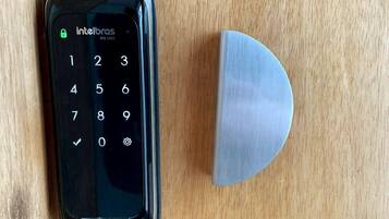 Um smartphone com teclado numérico e tela de bloqueio, apoiado em uma superfície de madeira ao lado de um objeto metálico.
