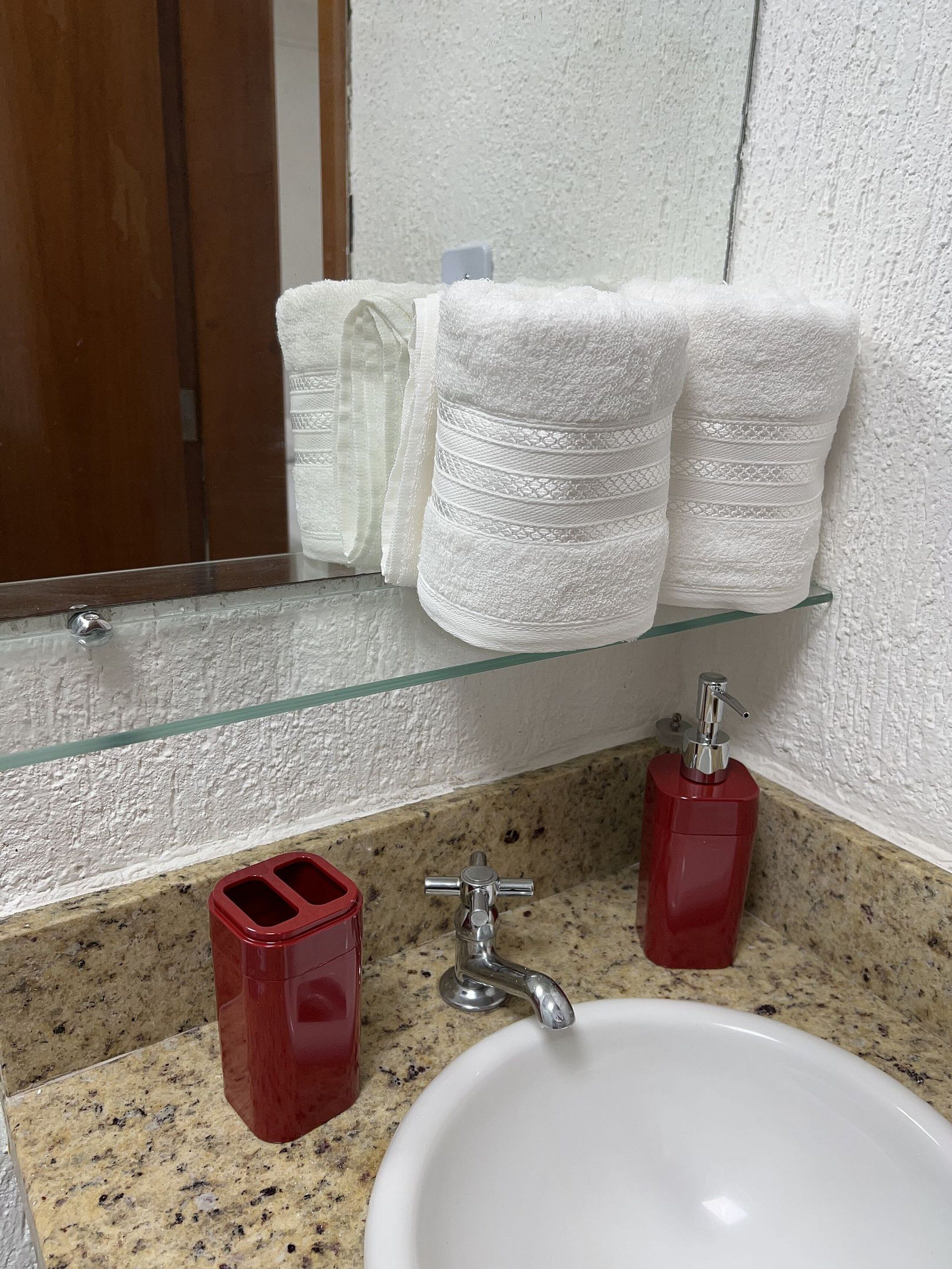 Um banheiro com bancada de granito, pia branca, prateleira de vidro com toalhas brancas e dois dosadores de sabonete vermelhos.