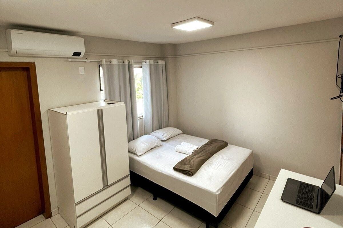 Un dormitorio con cama, escritorio con computadora portátil, aire acondicionado blanco y una puerta.