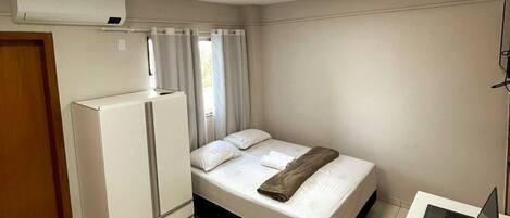 Un dormitorio con cama, escritorio con computadora portátil, aire acondicionado blanco y una puerta.
