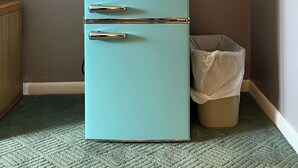 A teal Galanz refrigerator with two doors and a freezer compartment, placed in a corner with a beige carpet and a trash bin nearby. - Grand Lake Hotel (Lebanon)