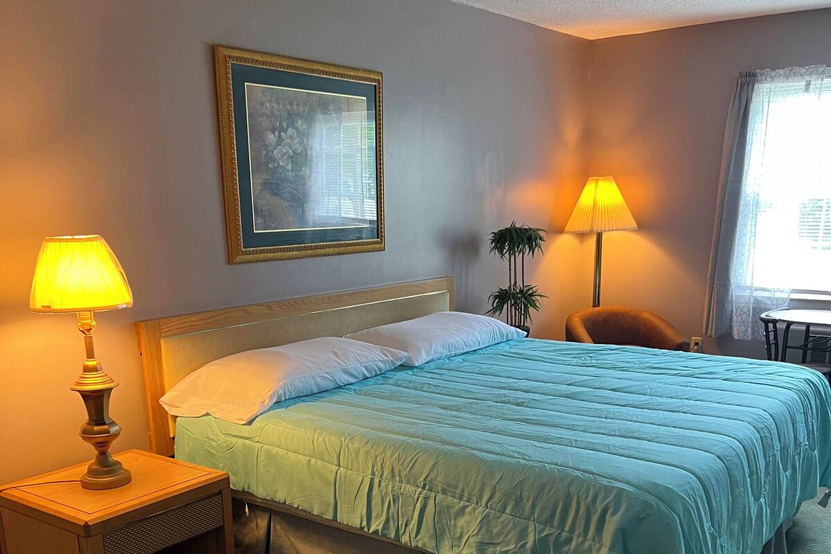 A hotel room with a bed, two bedside lamps, a framed picture on the wall, a chair, and a window with curtains.