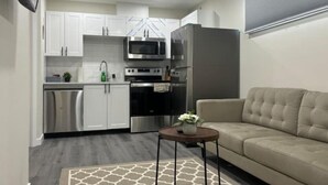 A compact kitchen with white cabinets, a microwave, oven, and refrigerator. There's a beige sofa and a small round table with a vase. - Guest Suite - Cozy Crib (Regina)