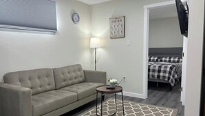 A modern living room with a grey sofa, a small side table, a patterned rug, a wall clock, a motivational poster, and a bed in the background. - Guest Suite - Cozy Crib (Regina)