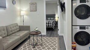 A compact living space with a sofa, a small table, a wall-mounted TV, a washing machine, and a bed with a checkered bedspread. - Guest Suite - Cozy Crib (Regina)