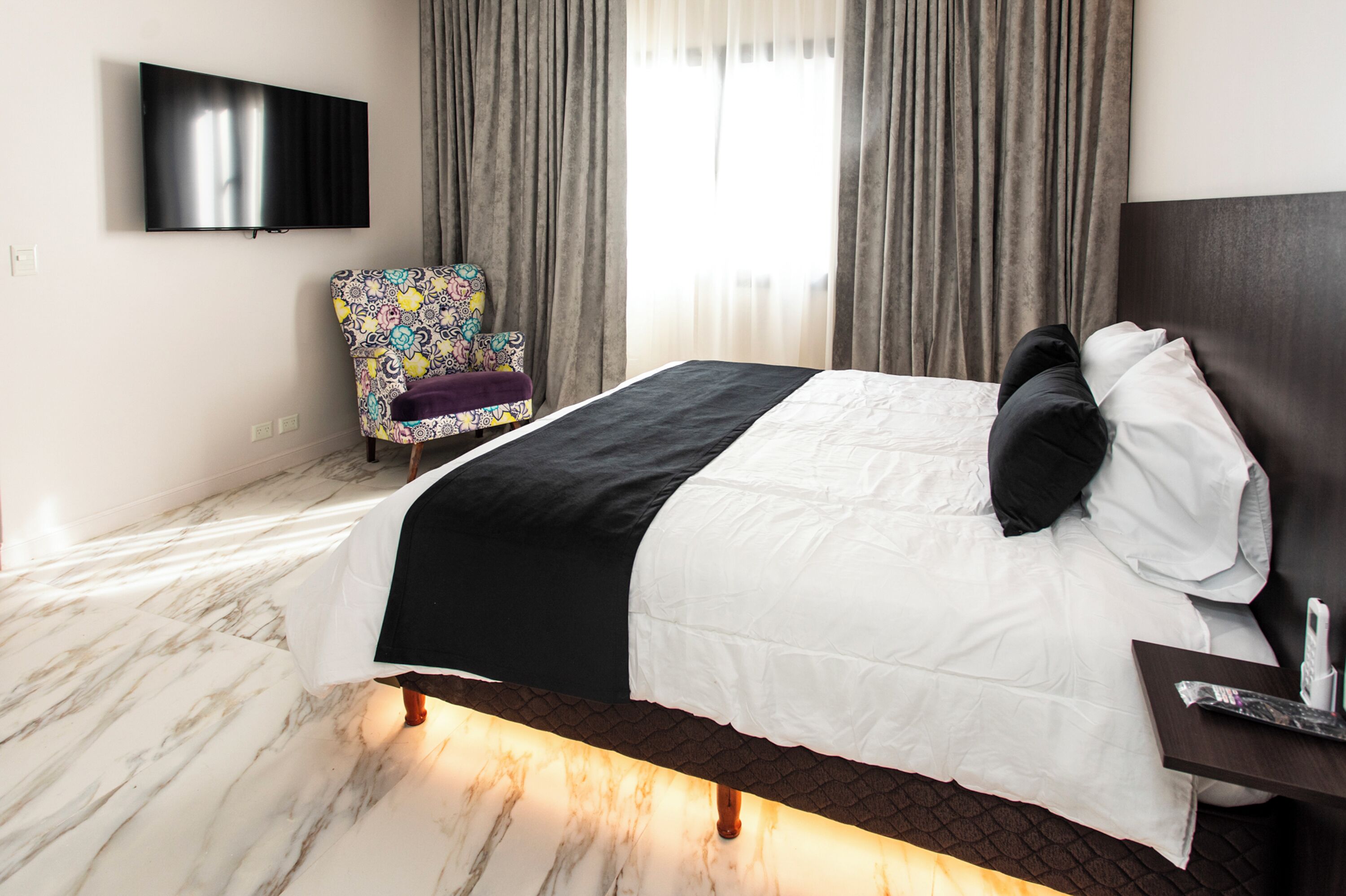 A hotel room with a bed, a TV mounted on the wall, a patterned armchair, and a window with curtains.