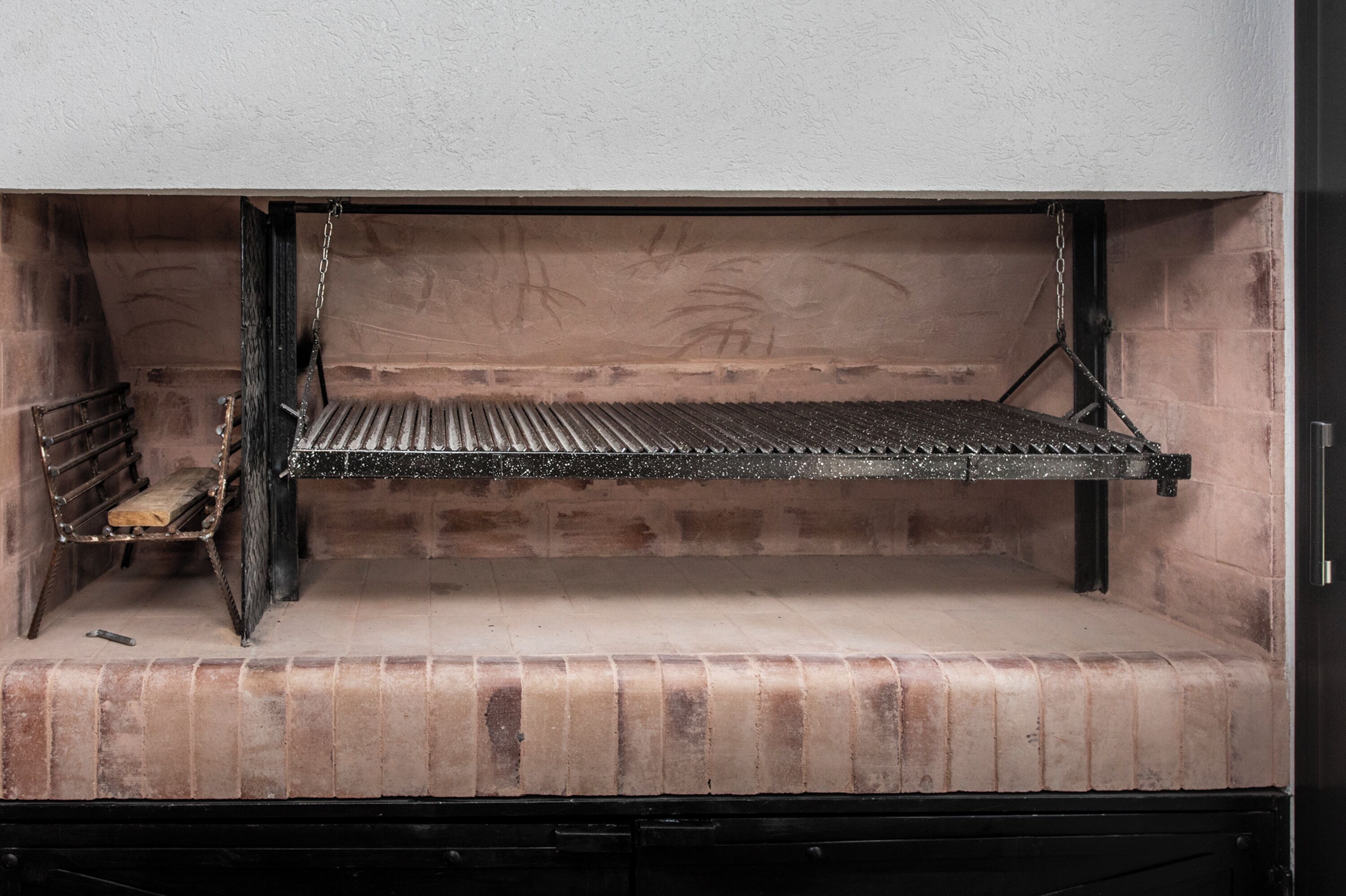 A fireplace with a metal grate, brick walls, and a metal shelf on the left.