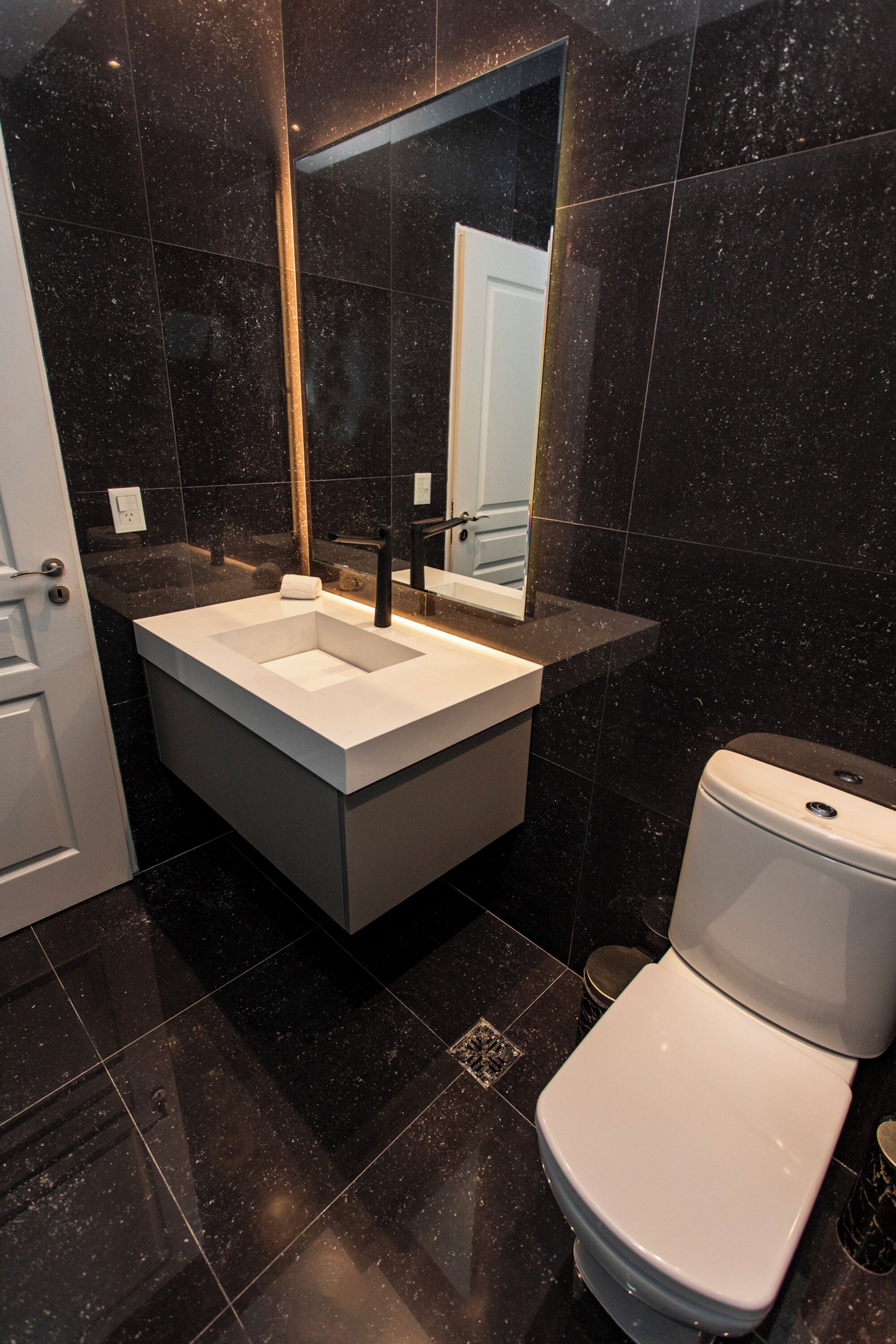 A modern bathroom with a white sink, black marble tiles, a white toilet, and a mirror above the sink.