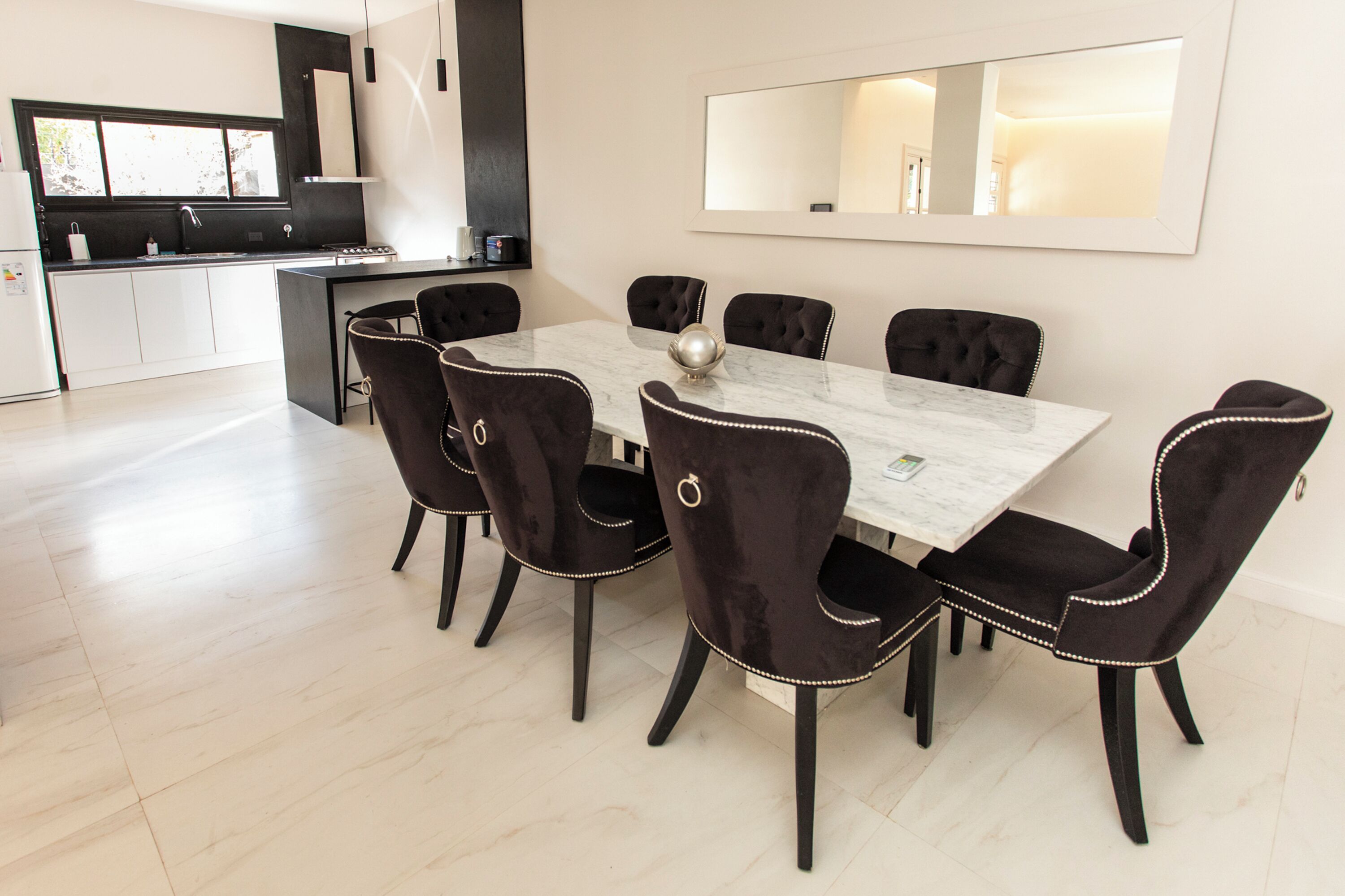 Un comedor moderno con una mesa de mármol y sillas tapizadas de color negro, una cocina al fondo y un espejo grande en la pared.