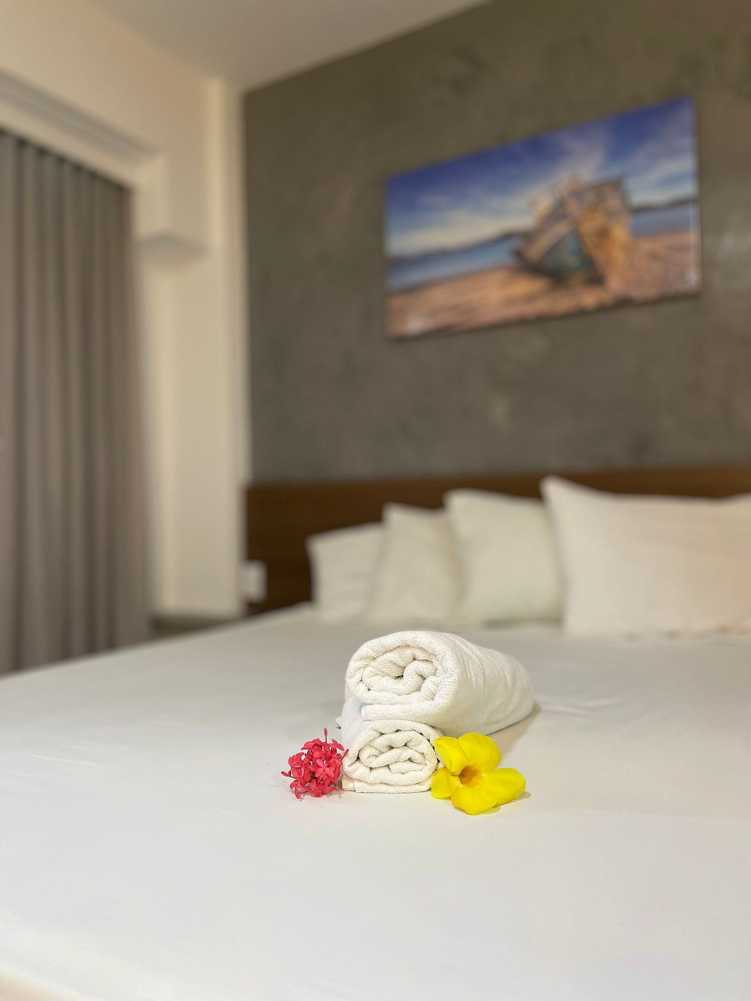A neatly made bed with a rolled white towel, a red flower, and a yellow flower on the bed.