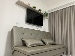 A modern living room with a grey sofa, two cushions, a mounted flat-screen TV, and a shelf with decorative items and plants. - Makia Beach Experience Porto de Galinhas (Ipojuca)