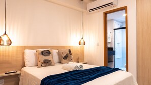 A hotel room with a bed, two bedside lamps, a wooden headboard, a blue blanket, and a view of the bathroom through an open door. - Makia Beach Experience Porto de Galinhas (Ipojuca)