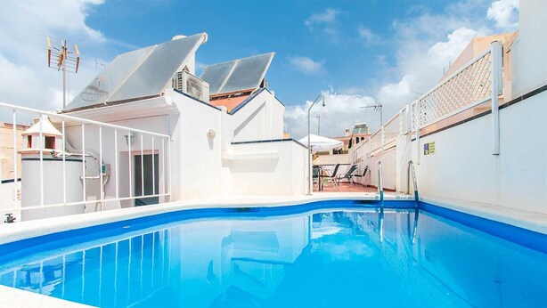 En swimmingpool med ett klart, blått vatten, omgiven av vita väggar och ett takförsett utomhusområde med stolar och parasoller.