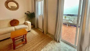 Sala de estar con sofá, una mesita de madera, una planta en maceta y vistas a un balcón con mobiliario exterior.