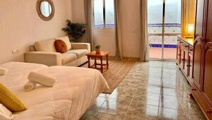 Habitación de hotel con cama, sofá, pequeña mesa con planta, espejo y vista a la playa a través de amplios ventanales.