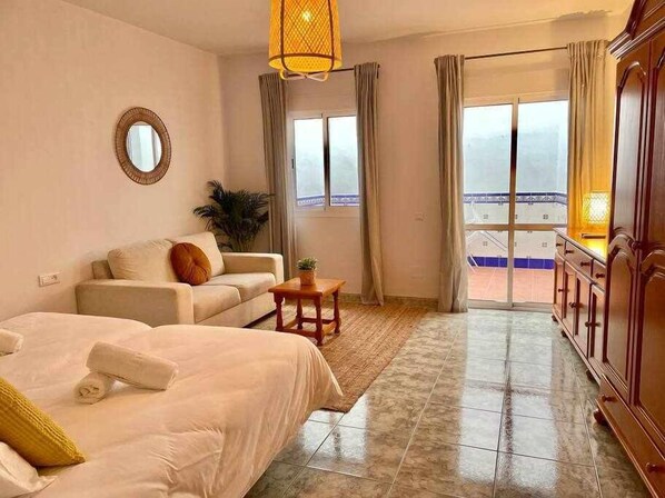 A hotel room with a bed, a sofa, a small table with a plant, a mirror, and a view of the beach through large windows.