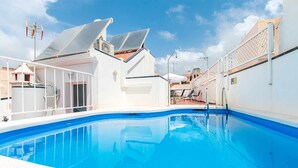 Una piscina con superficie de agua azul cristalina, rodeada de paredes blancas y un área exterior cubierta con sillas y sombrillas.
