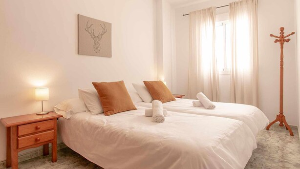 A hotel room with two beds, a bedside table, a lamp, a wooden floor, and a window with curtains.