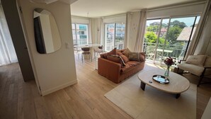 A spacious living room with a brown sofa, a coffee table, and a dining area with a round table and chairs. Large windows provide a view of the cityscape.