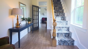 Un pasillo con una consola azul oscuro, una lámpara y una planta en maceta. Una escalera con alfombra estampada y balaustrada de madera conduce a otra habitación.