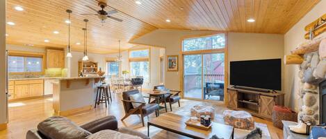 A spacious living room with a wooden ceiling, a stone fireplace, a television, and a kitchen area visible through large windows.