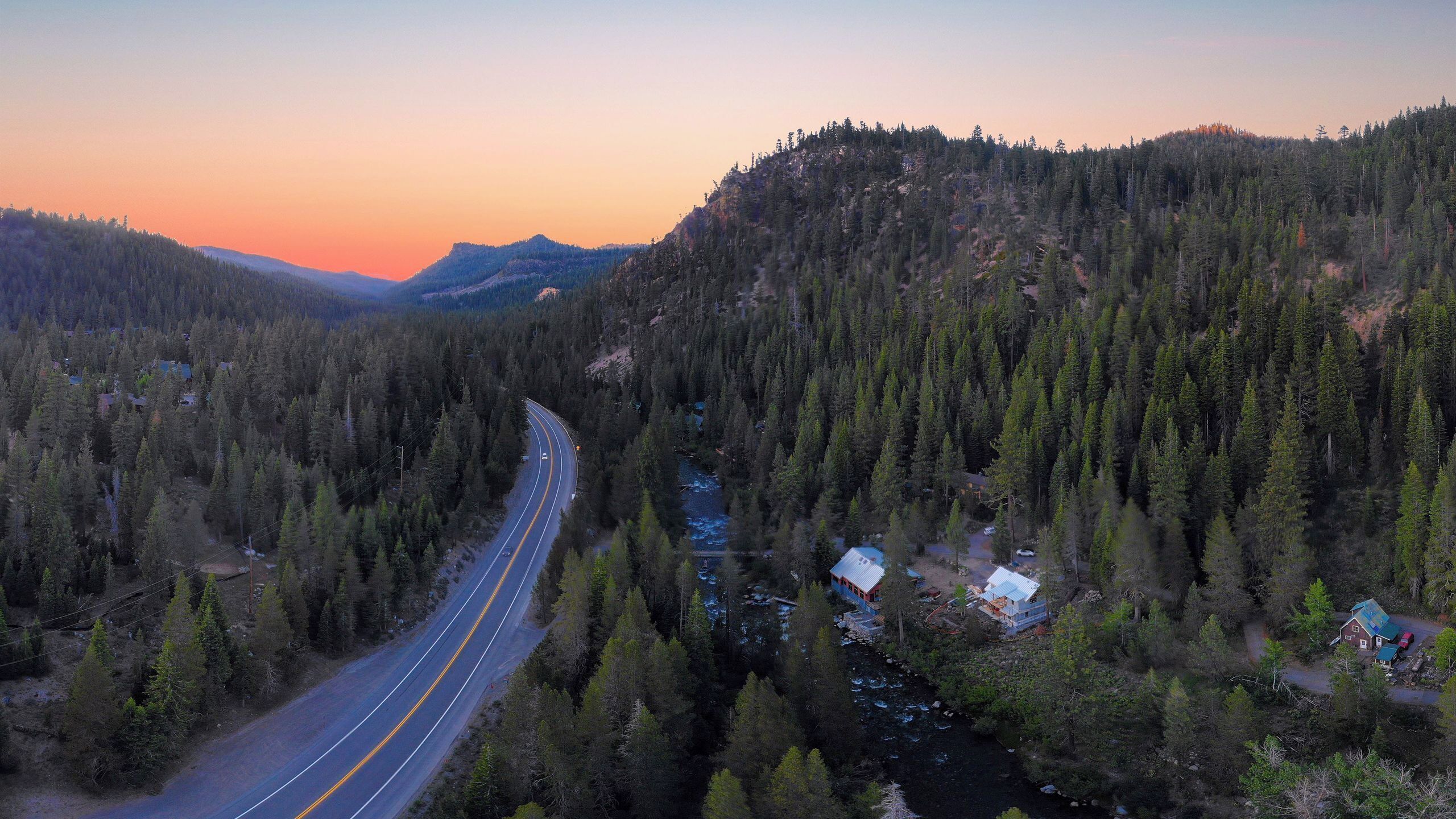 A winding road through a forested valley with a river, houses, and a mountain in the background.