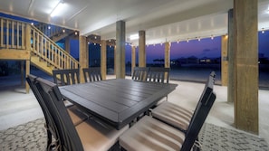 An outdoor patio with a wooden table and chairs, surrounded by wooden pillars, and a staircase leading to an upper level.