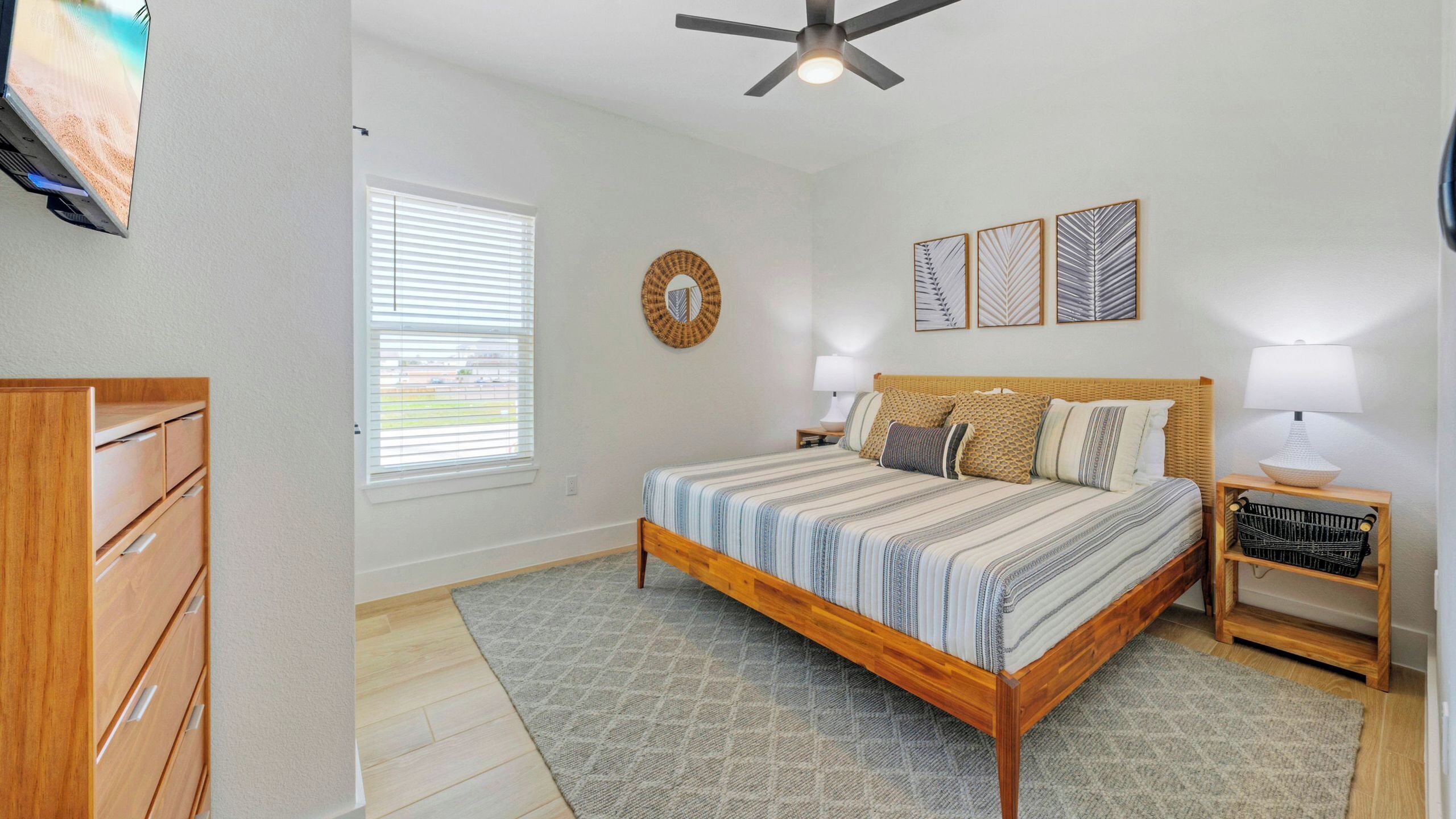 A bedroom with a bed, a nightstand, a lamp, a ceiling fan, a window with blinds, and a wall-mounted mirror.