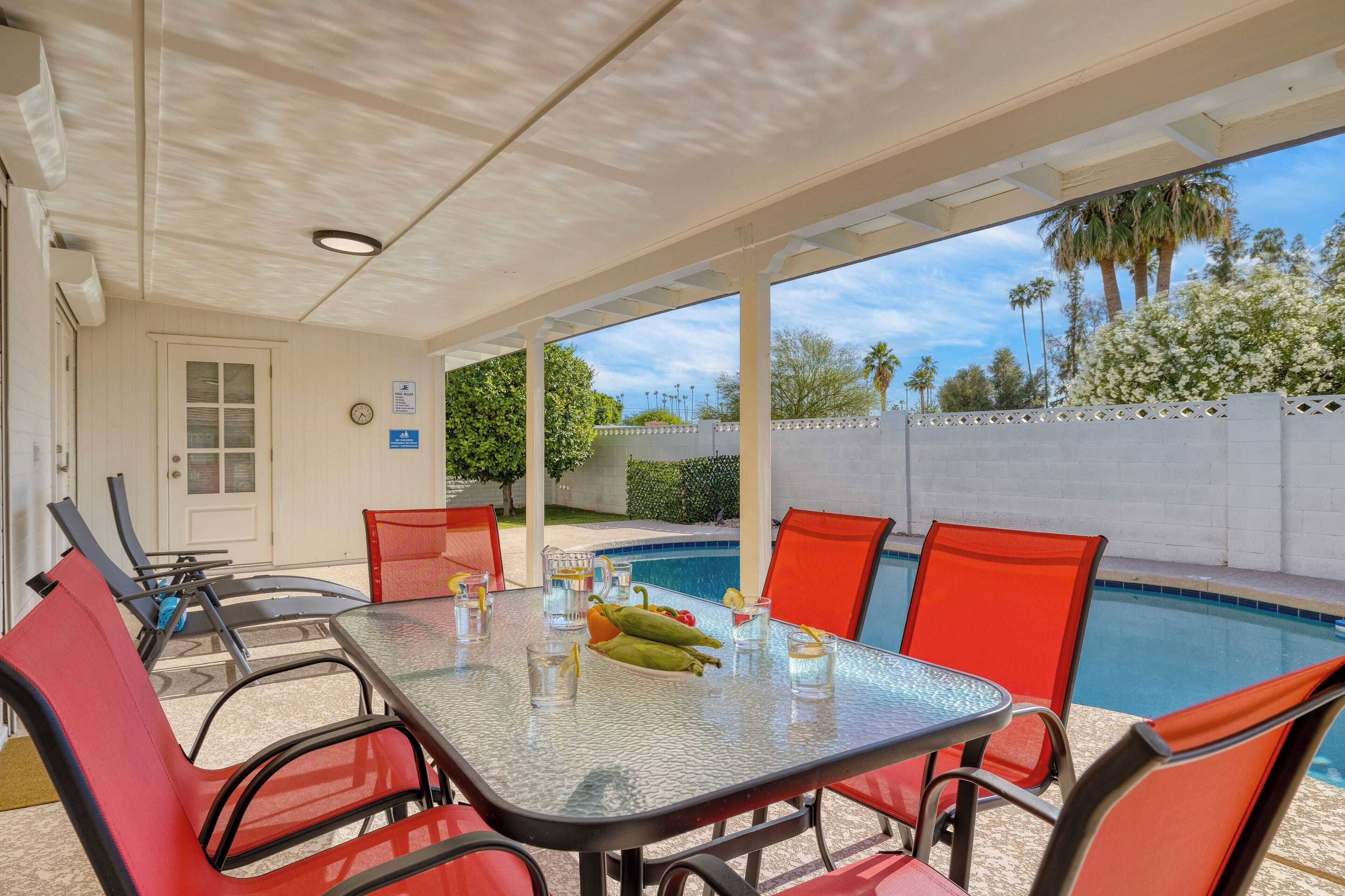 An outdoor patio with a glass table and red chairs, a pool, and a view of palm trees and a white fence.