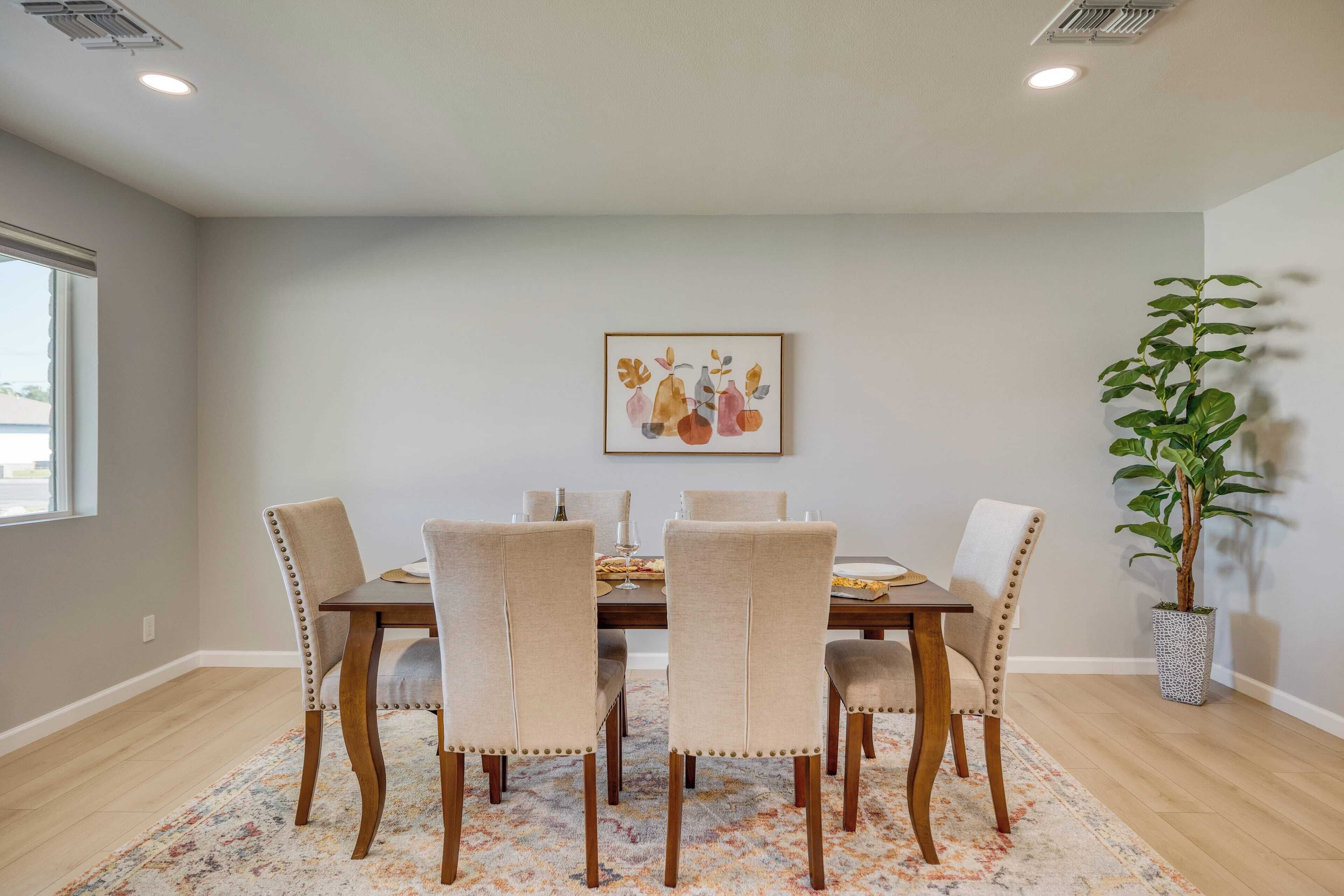 A dining area with a wooden table, beige upholstered chairs, a floral-patterned rug, a potted plant, and a framed artwork on the wall.