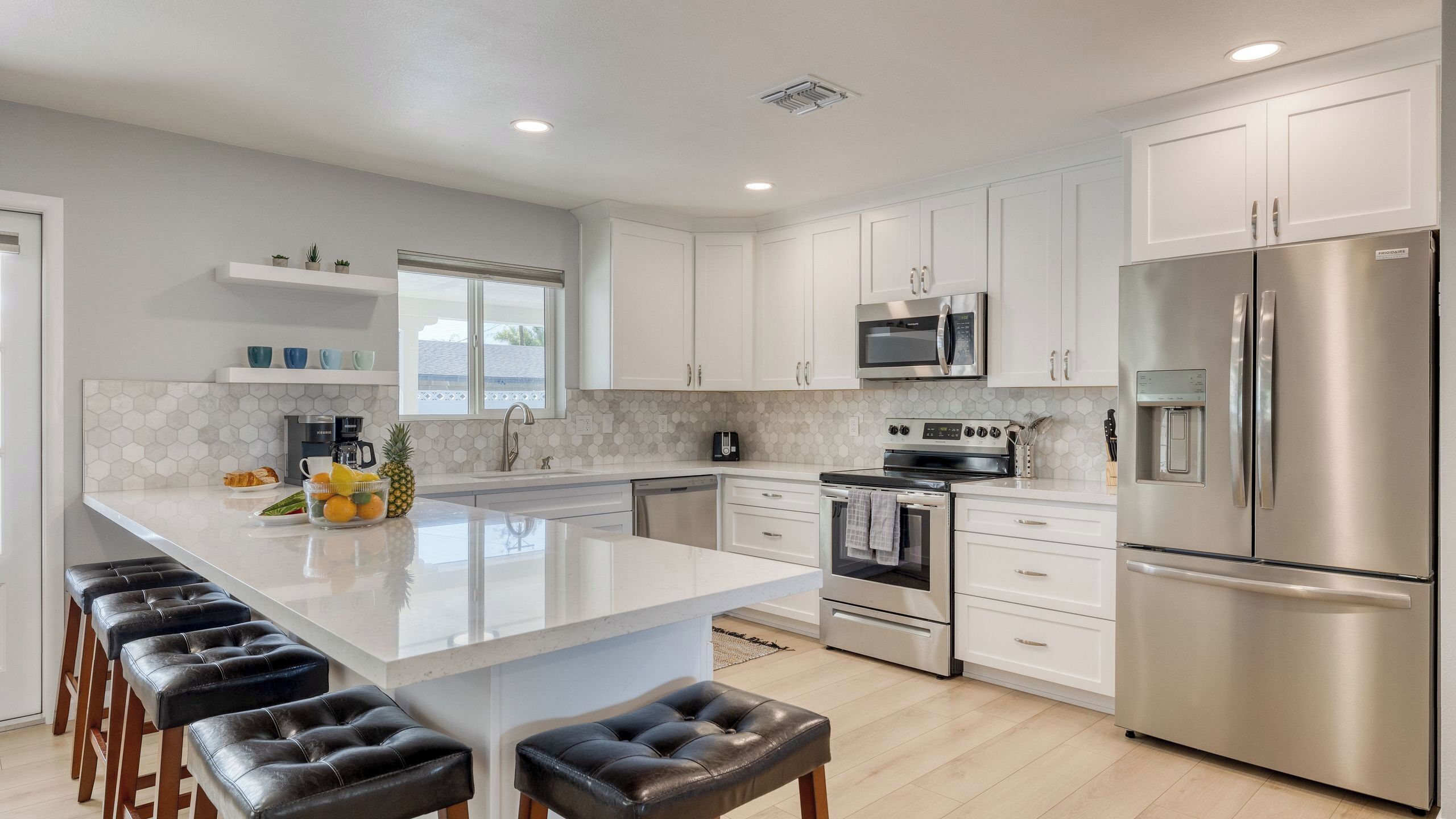 A modern kitchen with white cabinets, stainless steel appliances, a large island with bar stools, and a window providing natural light.