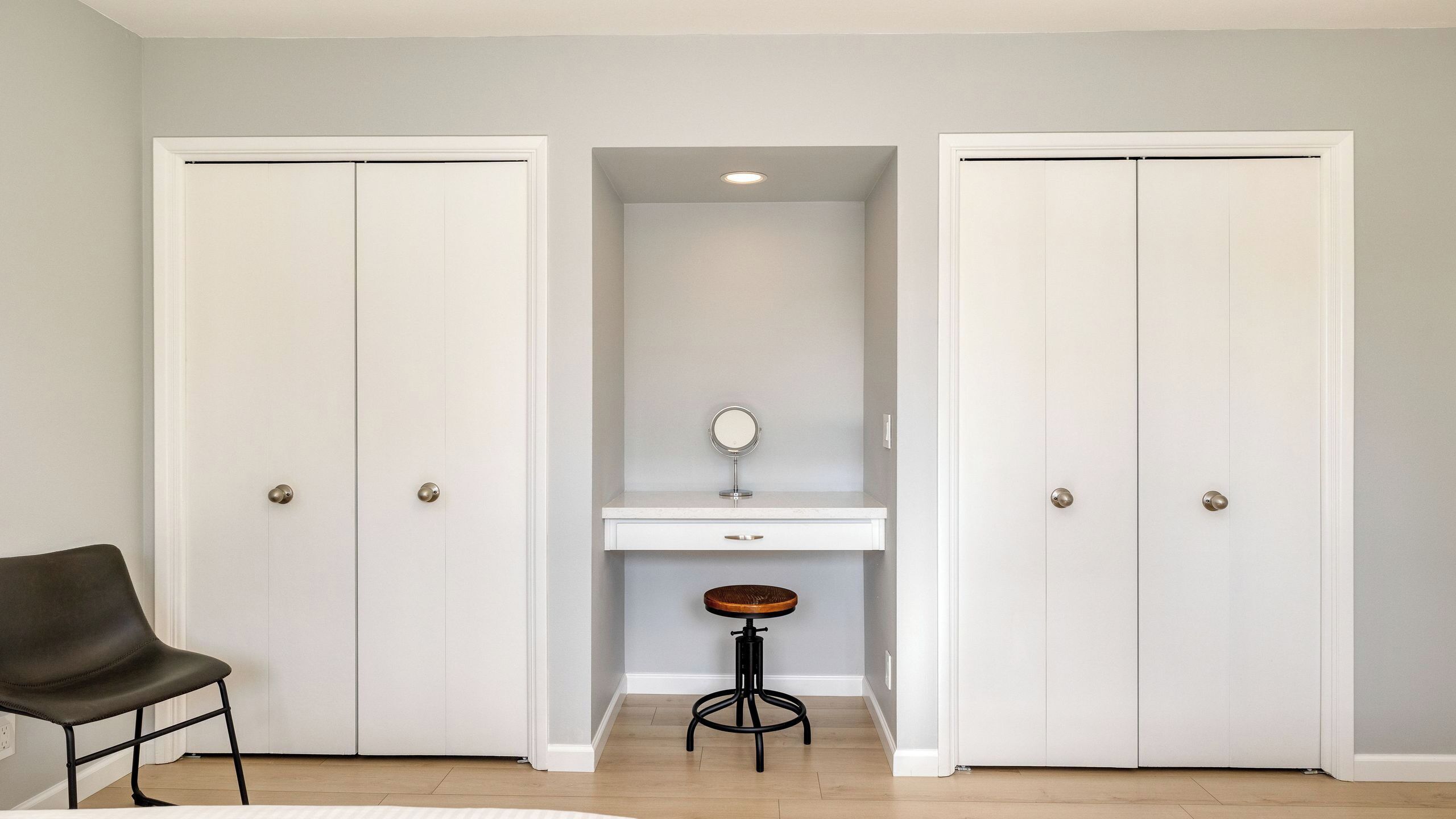 A room with two white doors, a small desk with a mirror, a stool, and a chair.
