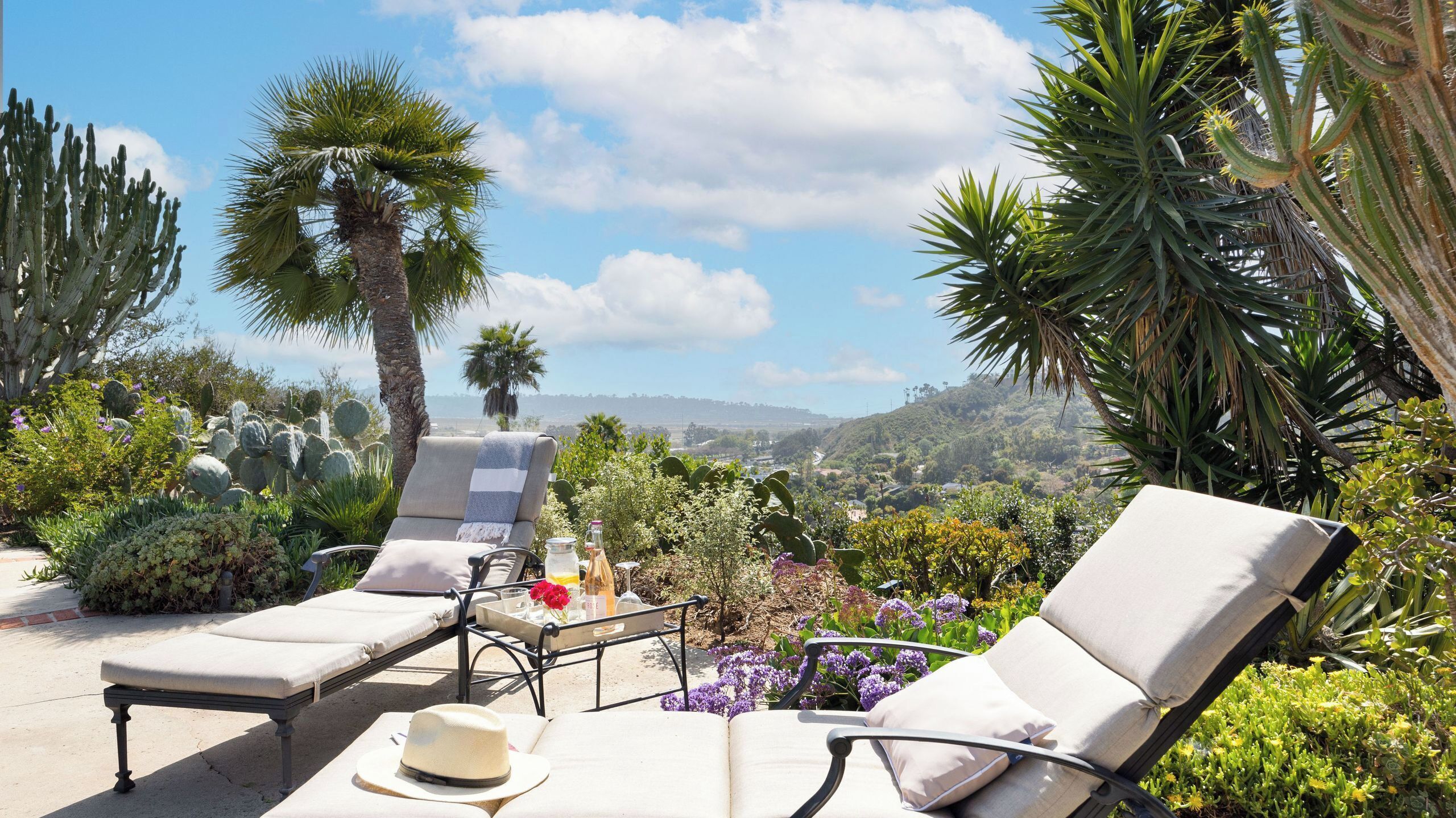A sunlit outdoor seating area with lounge chairs, a hat, and a bottle, surrounded by cacti and flowers.