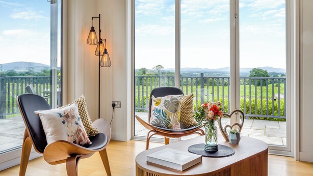 A spacious room with large windows, a wooden floor, a black chair with a patterned cushion, a round wooden table with a book and vase, and a decorative light fixture. - Whitehill Lodge - UK47194 (Walton, near Brampton)