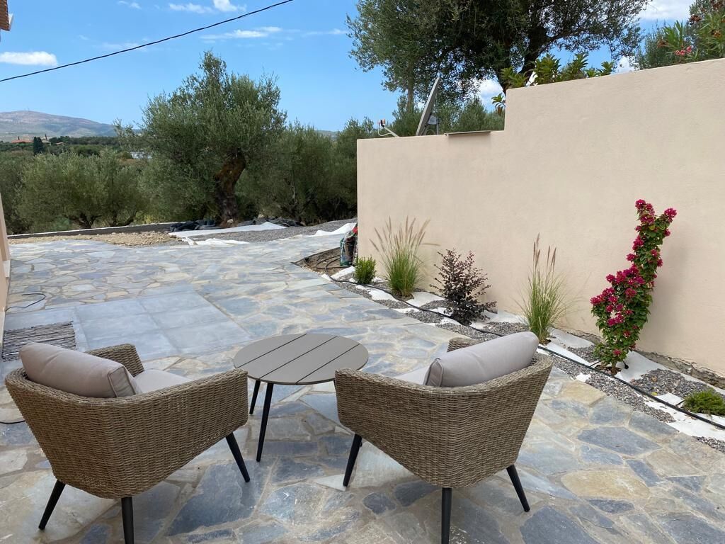 Un patio al aire libre con dos sillas de mimbre y una mesa, rodeado de suelo de piedra y plantas.