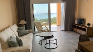 Una sala de estar con sofá, una mesa redonda, un televisor y vista al mar a través de amplios ventanales corredizos.