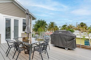 En terrasse med et bord med glasplade og stole, en overdækket grill og udsigt til palmer og en pool.