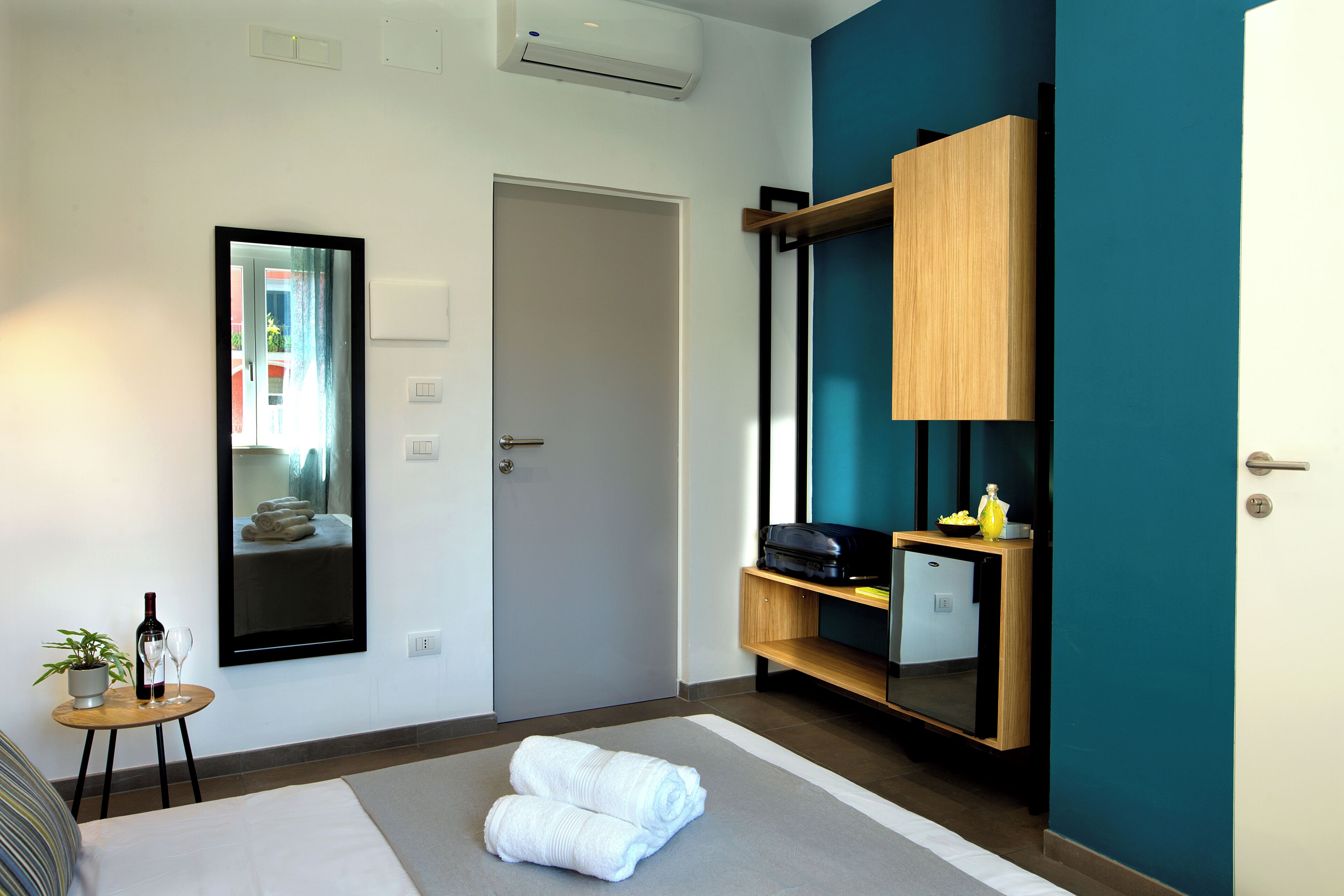 A hotel room with a bed, a small table with a plant and wine glasses, a mirror, a door, and a wooden shelf with a suitcase and a bowl.